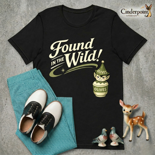 A top-down flat-lay photograph on a grey concrete surface featuring a black 'Found in the Wild!' Pixieware graphic tee. To the left is a pair of folded teal gingham checked pants with black and white saddle shoes. To the lower right are vintage ceramic knick-knacks: a fawn figurine and a winking olive jar pixie.