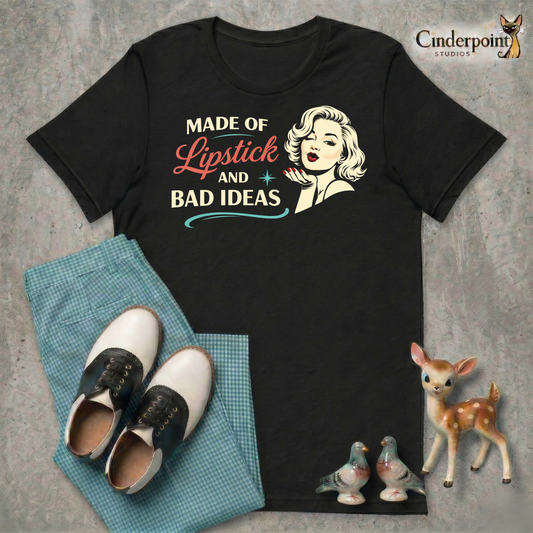 A top-down flat-lay on a grey concrete surface featuring the 'Lipstick and Bad Ideas' black tee. Included are teal gingham pants, black and white saddle shoes, and vintage ceramic figurines.