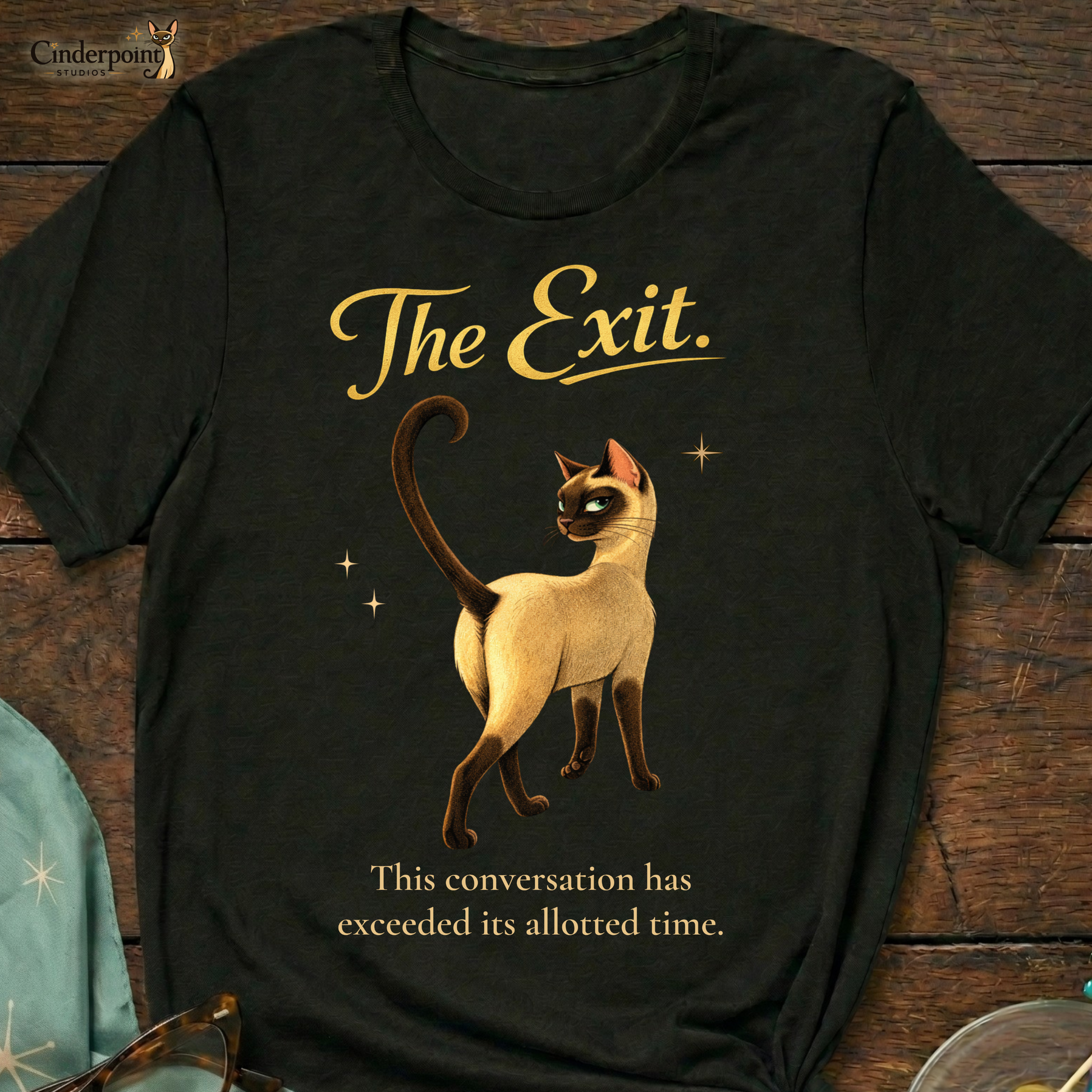 A black Bella+Canvas 3001 t-shirt featuring "The Exit" design on a dark wood grain table. The design shows a Siamese cat walking away with the text "The Exit" and "This conversation has exceeded its allotted time." Accessories include turquoise cat-eye glasses, a matching starburst scarf, and a glass dish with olives. Cinderpoint Studios logo in the corner.