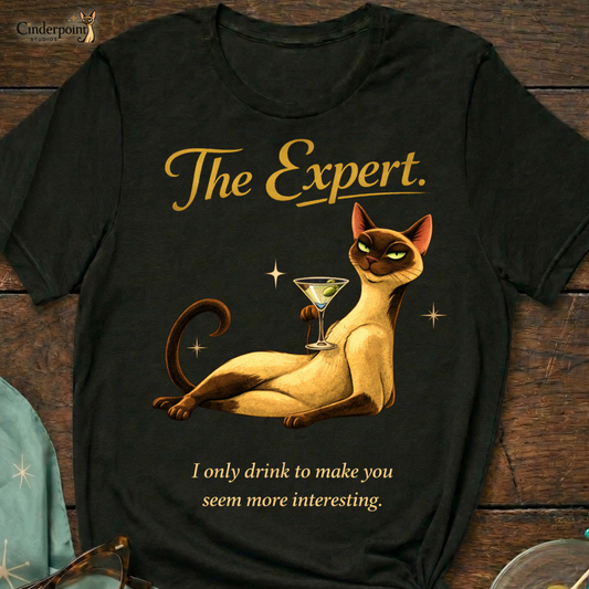 A black Bella+Canvas 3001 t-shirt featuring "The Expert" design on a dark wood grain table. The design shows a Siamese cat lounging with a martini and the text "The Expert" and "I only drink to make you seem more interesting." Accessories include vintage cat-eye glasses and a cocktail garnish dish. Cinderpoint Studios logo in the corner.
