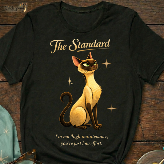 A black Bella+Canvas 3001 t-shirt featuring "The Standard" design, placed on a dark wood grain table. The design shows a stylized Siamese cat, the text "The Standard," and the quote "I’m not high maintenance. You’re just low effort." Next to the shirt are accessories: turquoise cat-eye glasses, a matching turquoise atomic starburst scarf, and a small glass dish with green olives on decorative vintage cocktail picks. The Cinderpoint Studios logo is in the top left.