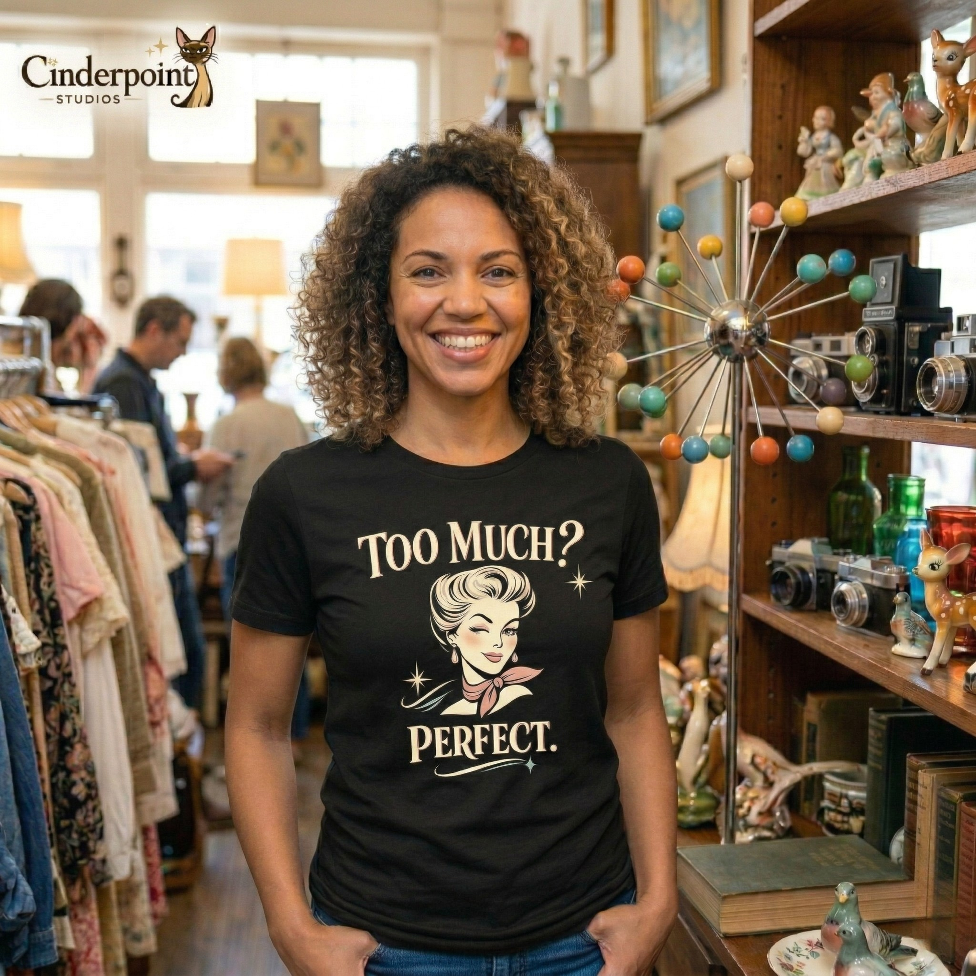 A medium shot of a cheerful woman with curly brown hair wearing the 'Too Much? Perfect.' black tee. She is standing in a vintage store near a prominent atomic-age lamp and wooden shelves of curios.
