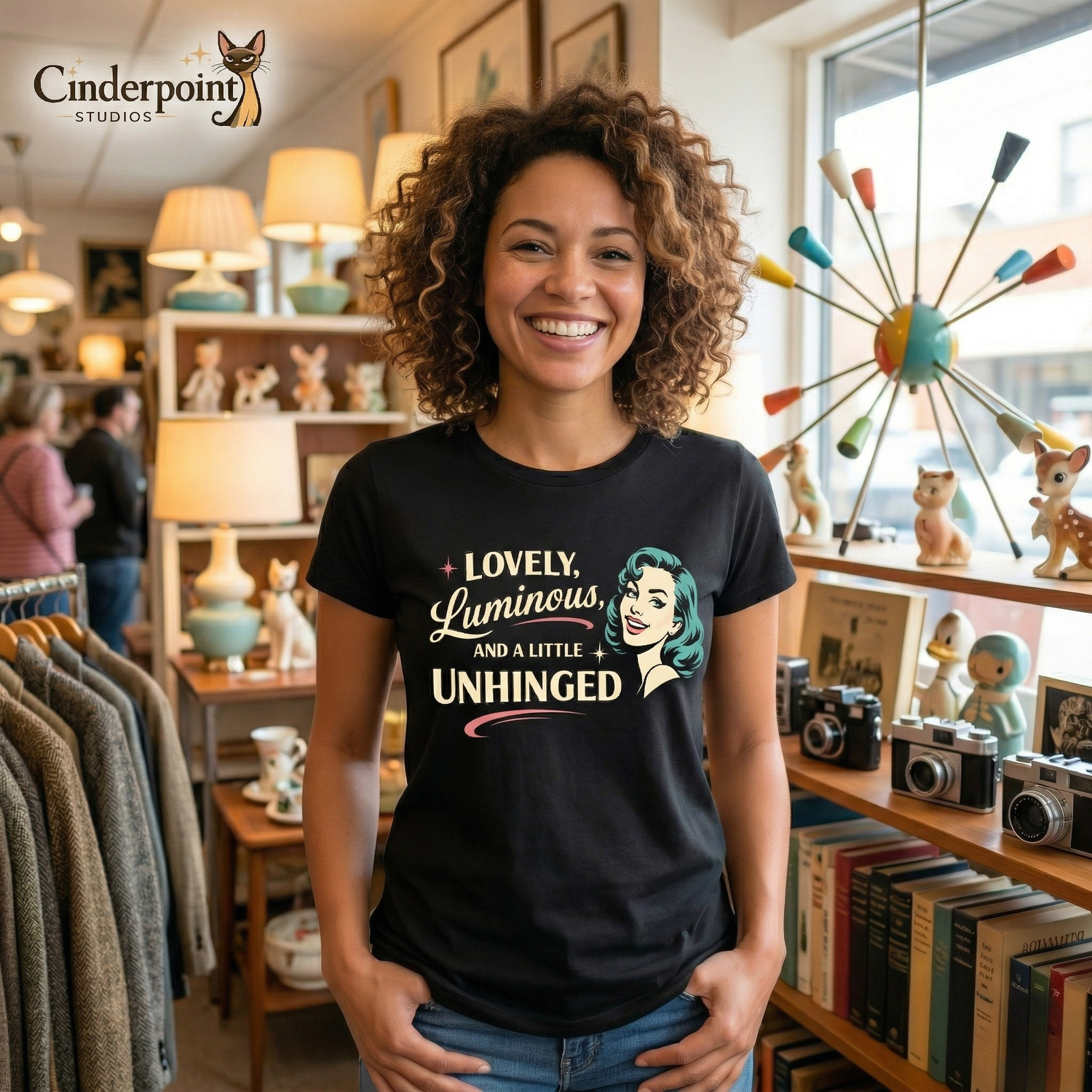 A medium shot of a cheerful woman with curly brown hair wearing the 'Lovely Luminous' black tee. She is standing in a vintage store near a prominent atomic-age lamp and wooden shelves of curios.