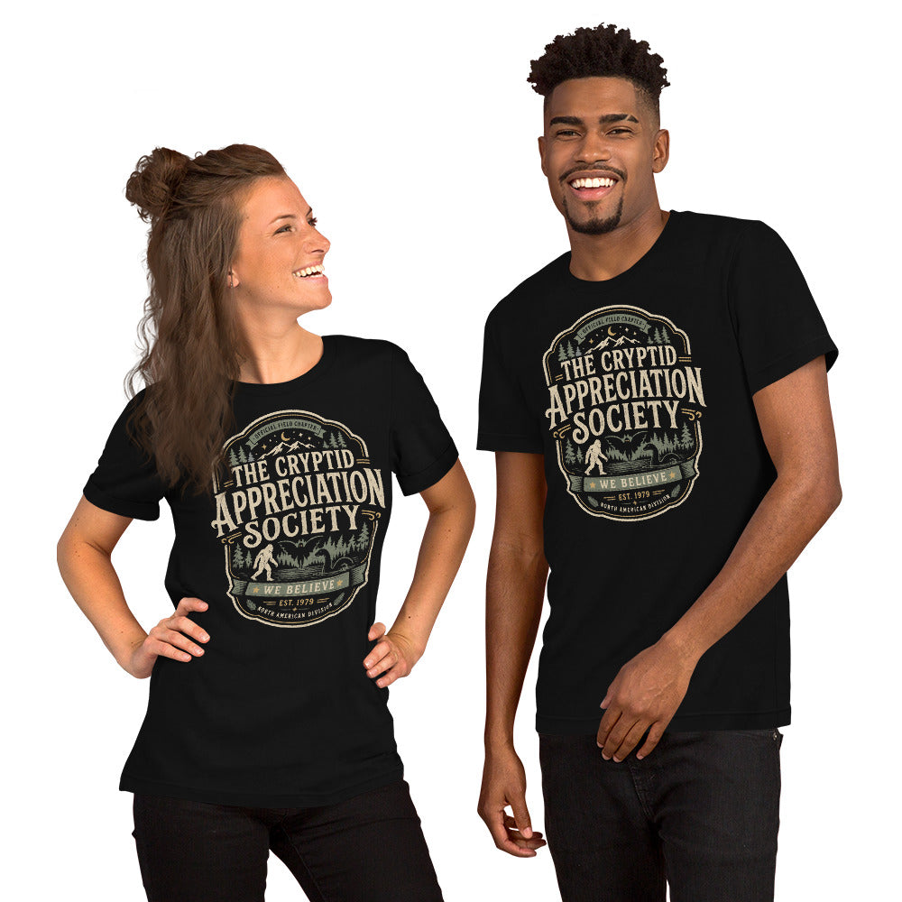 Man & woman wearing a black graphic tee with The Cryptid Appreciation Society artwork by Cinderpoint Studios, styled as a boutique retro paranormal and cryptid fan shirt with Sasquatch, Mothman, Nessie-style creature, and forest badge artwork.