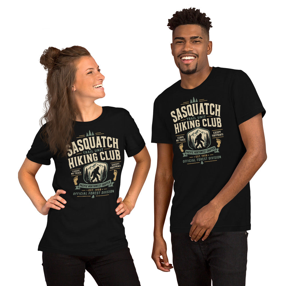 Black graphic tee featuring original Cinderpoint Studios artwork for Sasquatch Hiking Club, a retro Bigfoot trail design with bold varsity lettering, mountain scenery, pine trees, footprint accents, and text including Trail Unit, Pacific Northwest Chapter, Leave No Trace, Except Footprints, and Official Forest Division.