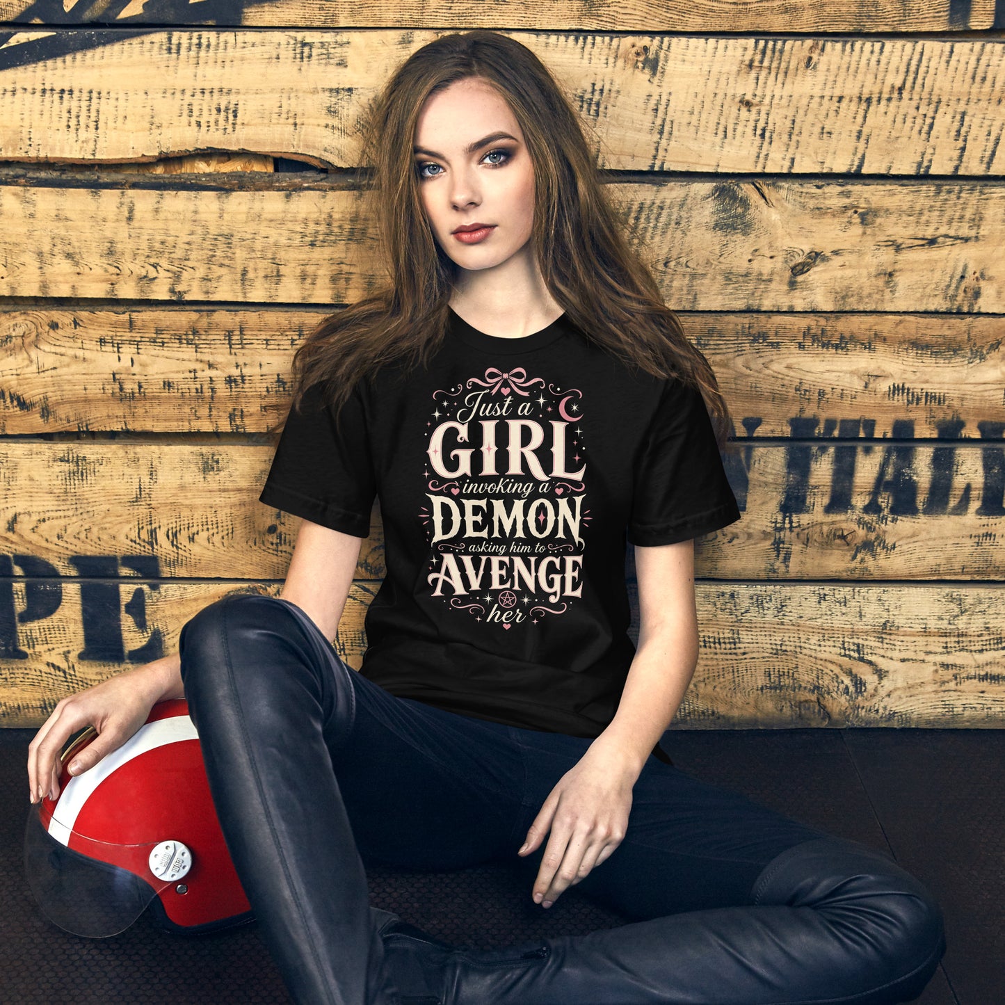 A young woman wearing a black graphic tee featuring original Cinderpoint Studios artwork for Just a Girl Invoking a Demon, a feminine occult humor design with bold vintage lettering, pink bow accents, crescent moon, stars, hearts, and a small pentagram.
