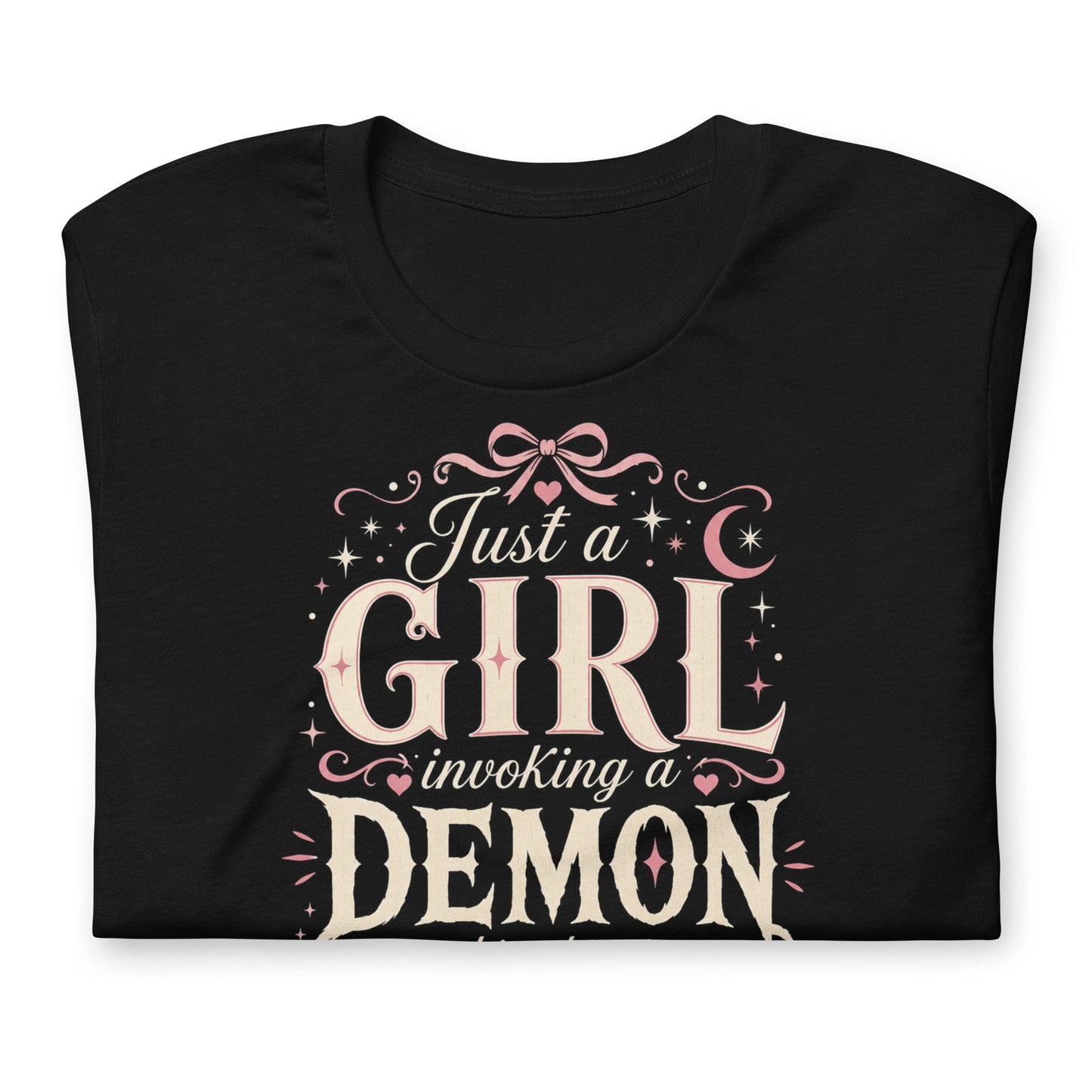 Black graphic tee featuring original Cinderpoint Studios artwork for Just a Girl Invoking a Demon, a feminine occult humor design with bold vintage lettering, pink bow accents, crescent moon, stars, hearts, and a small pentagram.
