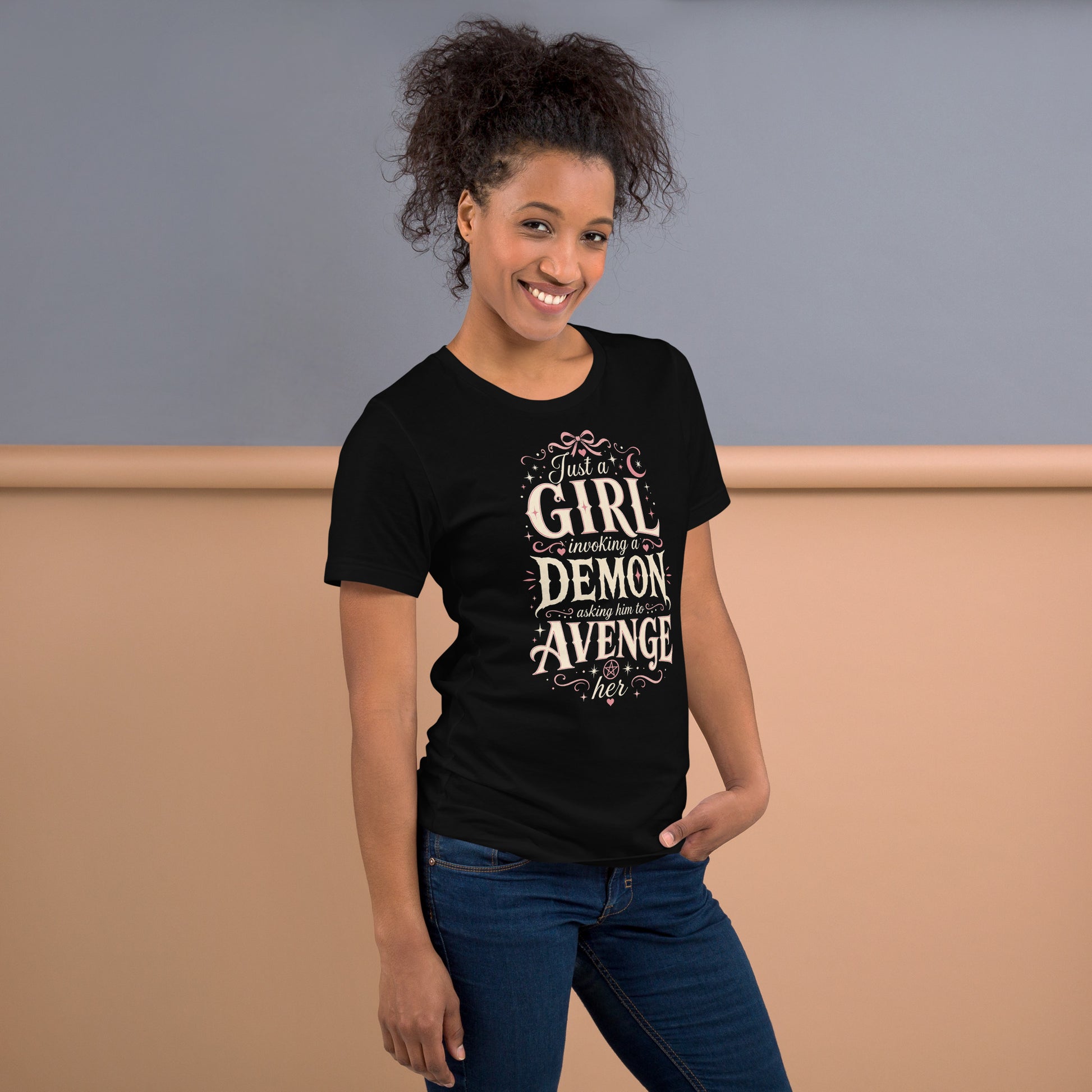 A pretty woman wearing a black graphic tee featuring original Cinderpoint Studios artwork for Just a Girl Invoking a Demon, a feminine occult humor design with bold vintage lettering, pink bow accents, crescent moon, stars, hearts, and a small pentagram.