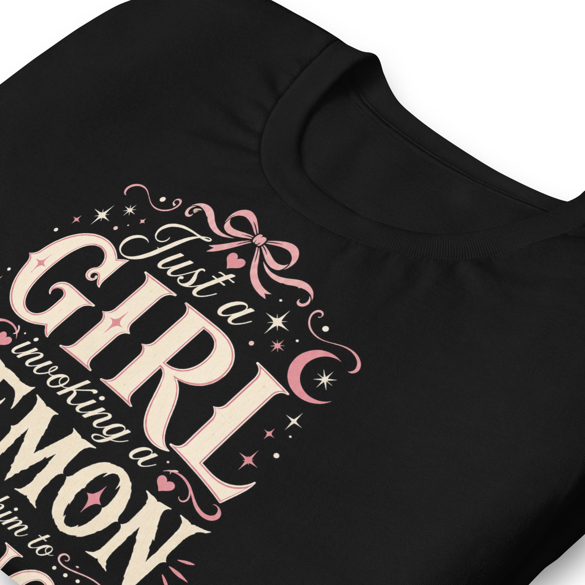 Black graphic tee featuring original Cinderpoint Studios artwork for Just a Girl Invoking a Demon, a feminine occult humor design with bold vintage lettering, pink bow accents, crescent moon, stars, hearts, and a small pentagram.